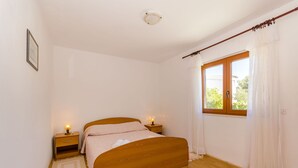 A bedroom with a wooden bed, two bedside tables with lamps, a window with curtains, and a white wall. - Apartment Lavcevic (Vrboska)