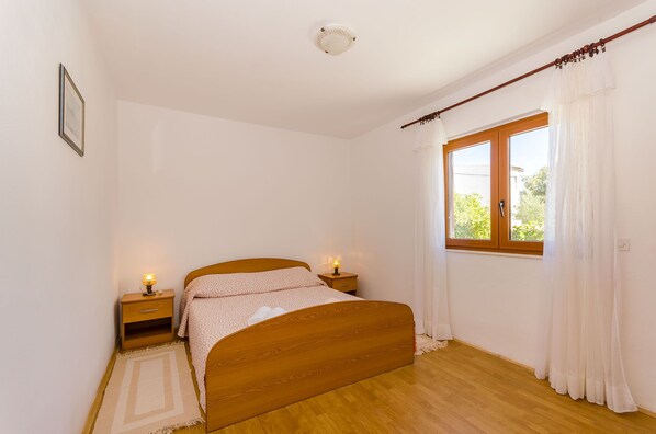 A bedroom with a wooden bed, two bedside tables with lamps, a window with curtains, and a framed picture on the wall. - Apartment Lavcevic (Vrboska)