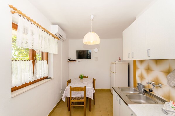 A small, clean kitchen with a dining table, a hanging light fixture, and a window with curtains. - Apartment Lavcevic (Vrboska)