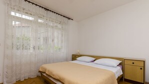 A bedroom with a bed, a nightstand, a wooden floor, and a window with sheer curtains. - Apartment Tome G (Primosten)