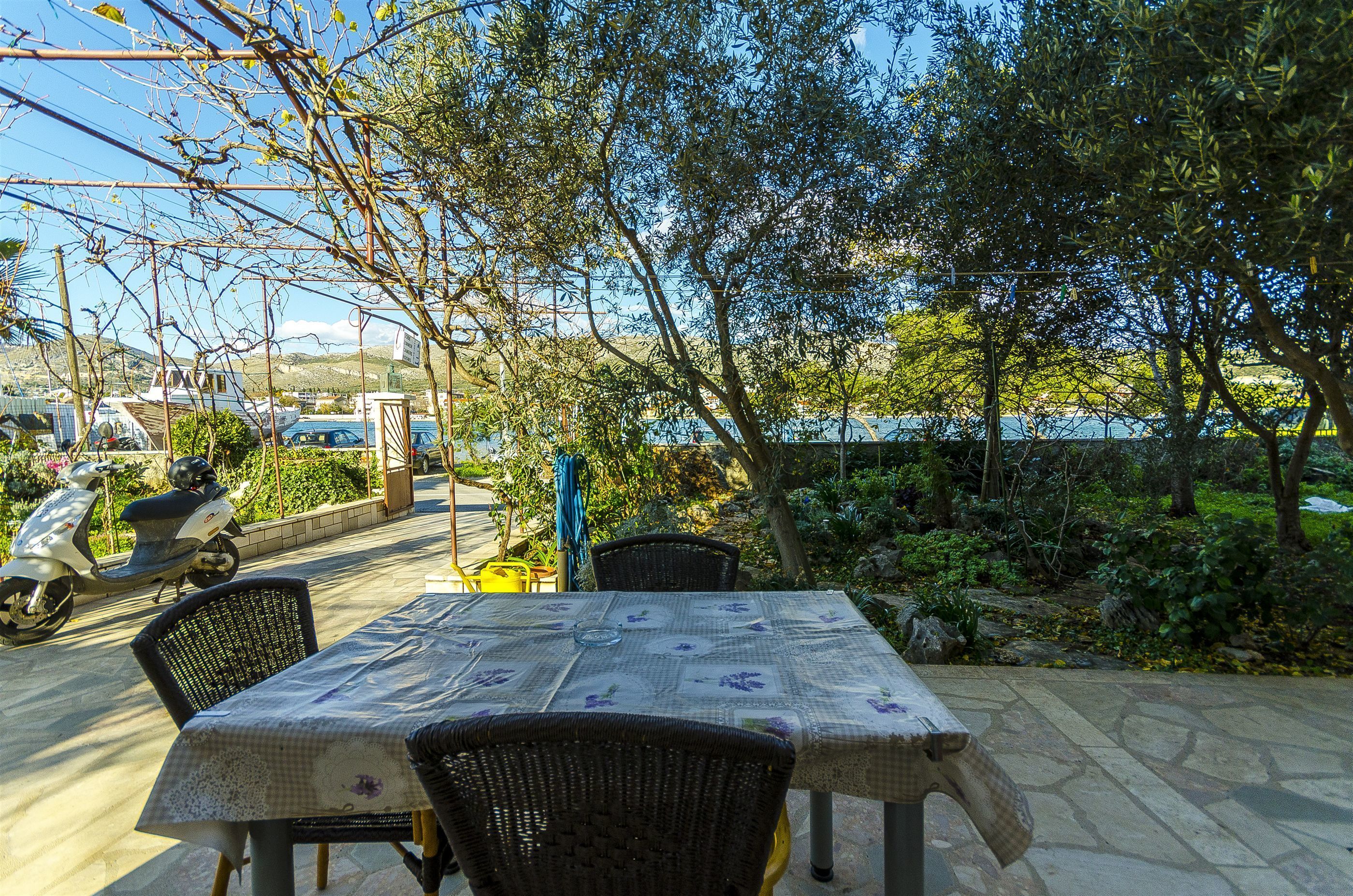 En udendørs patio med et bord og stole, omgivet af grøn vegetation med udsigt over vand.