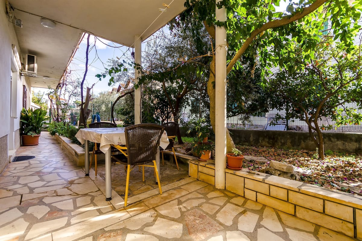 A covered patio with a stone floor, a table, and chairs, surrounded by potted plants and trees.
