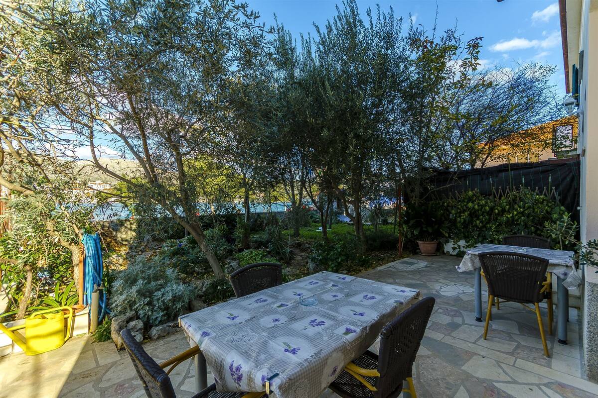 An outdoor patio with a table set for dining, surrounded by greenery and a view of the sea.