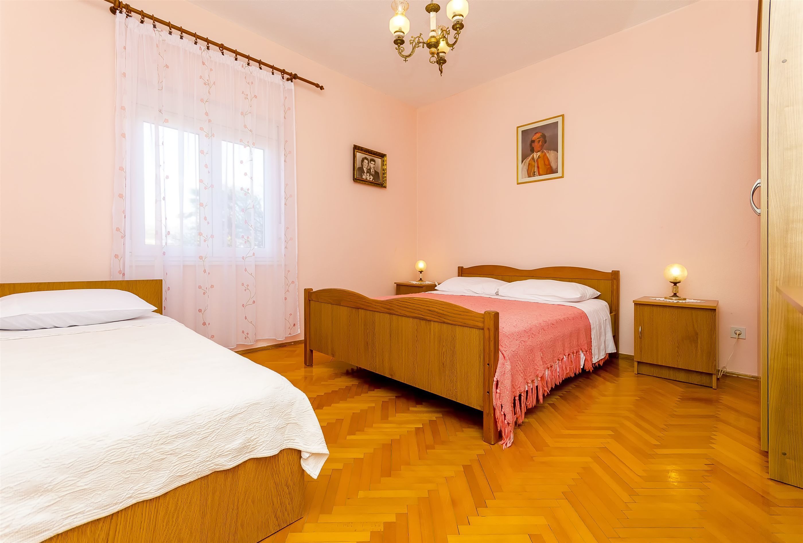 A bedroom with two wooden beds, a wooden nightstand, a bedside table, a chandelier, and a window with sheer curtains.