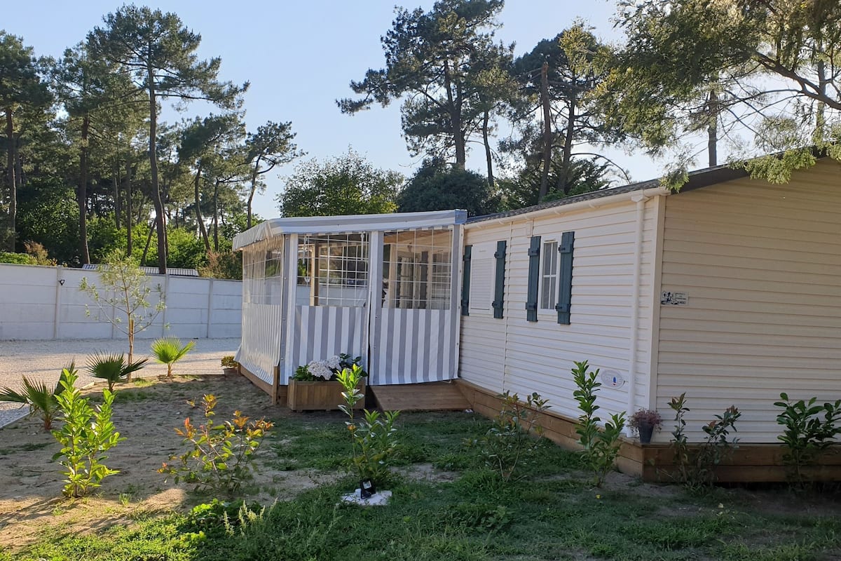 Ein Mobilheim mit überdachter Veranda, umgeben von einem eingezäunten Garten mit verschiedenen Pflanzen und Bäumen.