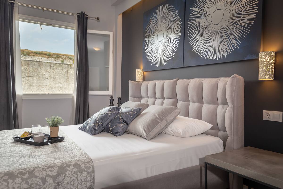 A modern bedroom with a tufted headboard, a bedside table, a tray with breakfast items, and a view of the outdoors through a window.