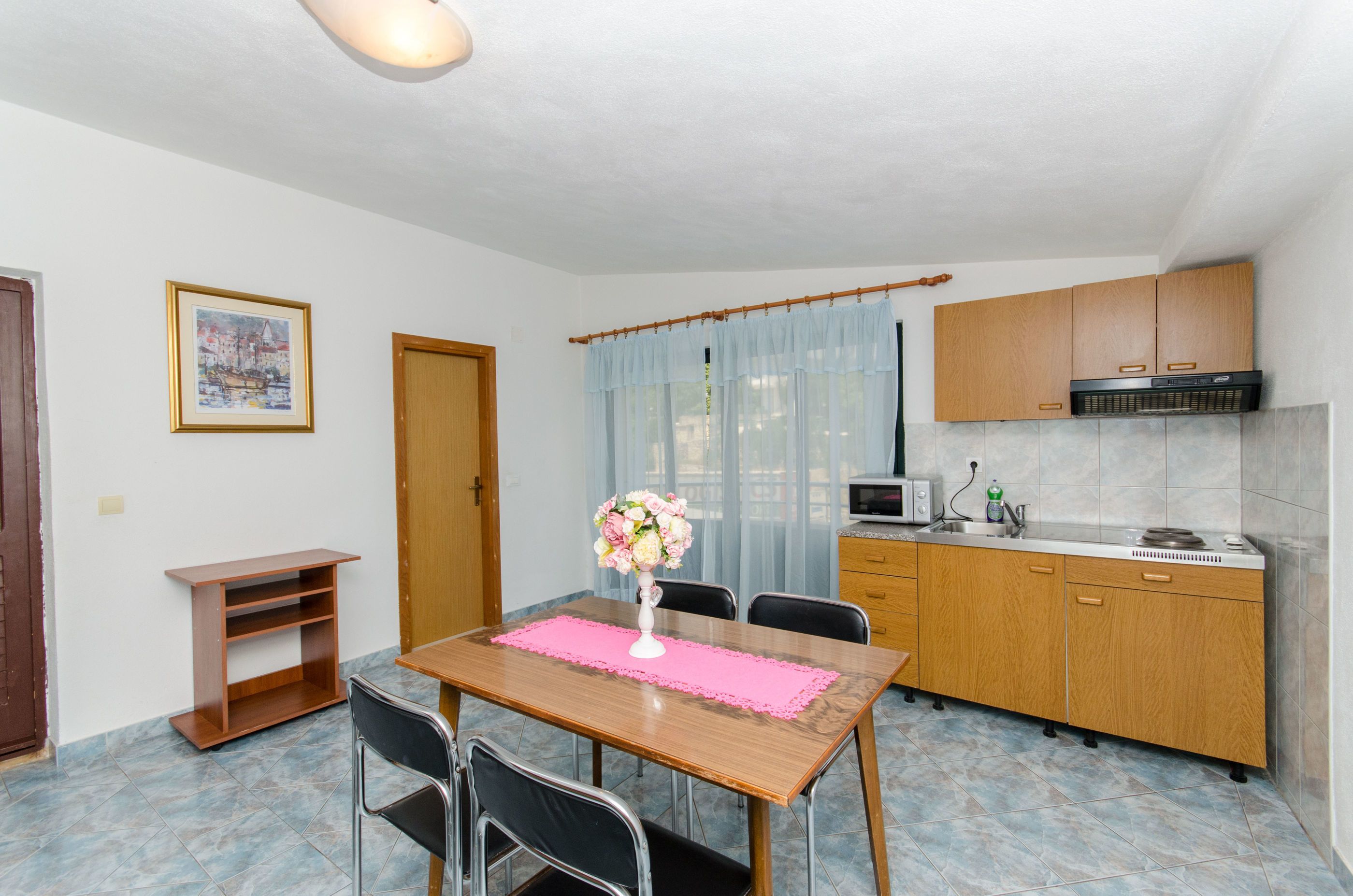 A compact kitchen with wooden cabinets, a microwave, and a sink. There's a dining table with chairs and a pink placemat. A framed picture hangs on the wall, and there's a window with sheer curtains.