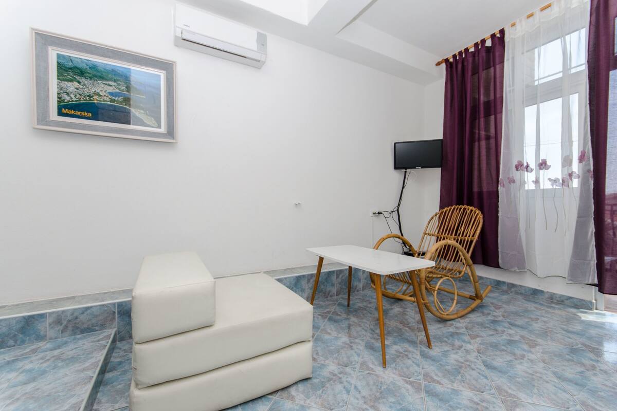 Ein Zimmer mit einem weißen Sofa, einem kleinen Tisch, einem Korbstuhl, einem Fernseher und einem großen Fenster mit violetten Vorhängen.