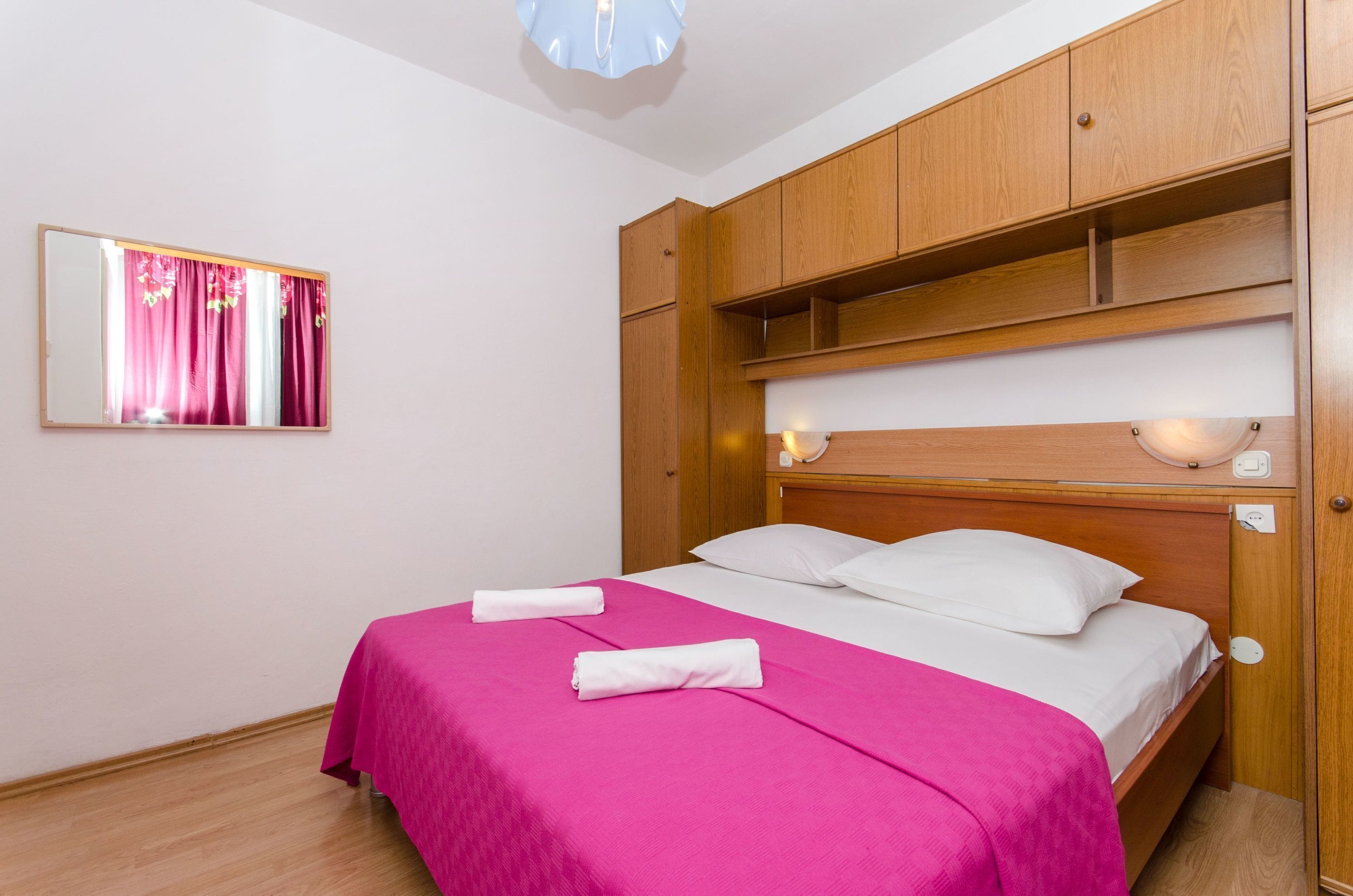 Une chambre d’hôtel avec un lit en bois, du linge de lit blanc, une couvre-lit rose et un miroir avec des rideaux.