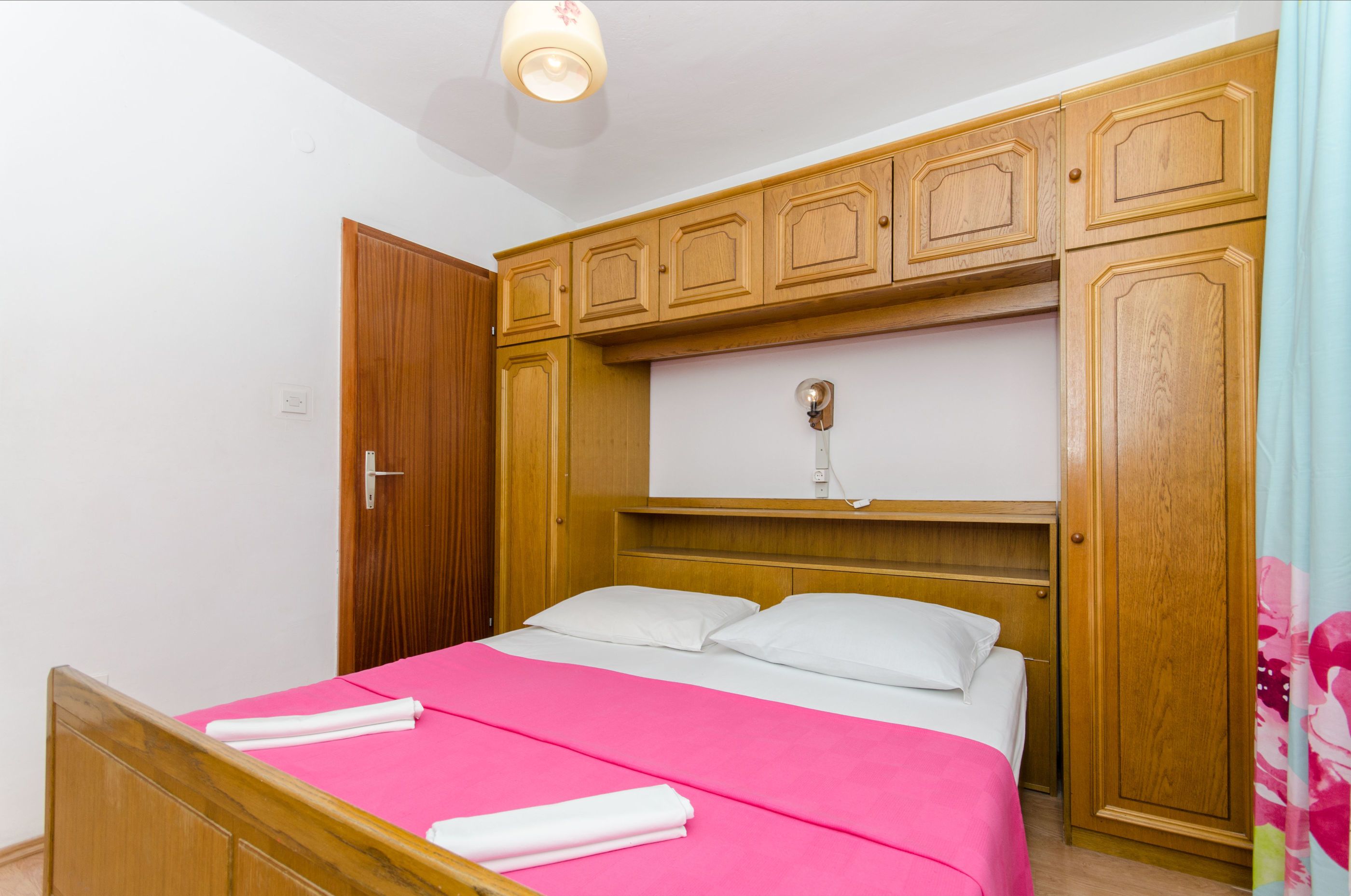 A hotel room with a wooden bed frame, a pink bedspread, white pillows, and wooden wardrobe cabinets.