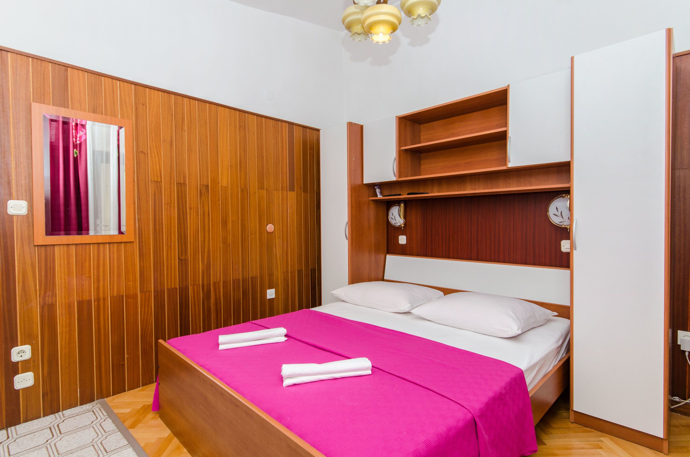 A hotel room with a wooden wardrobe, a bed with a pink bedspread, and a small shelf above the bed.