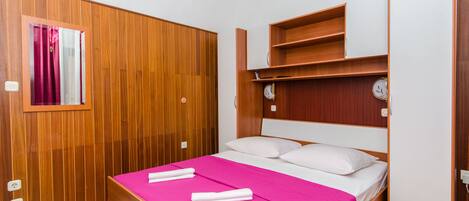A hotel room with a wooden wardrobe, a bed with a pink bedspread, and a small shelf above the bed.
