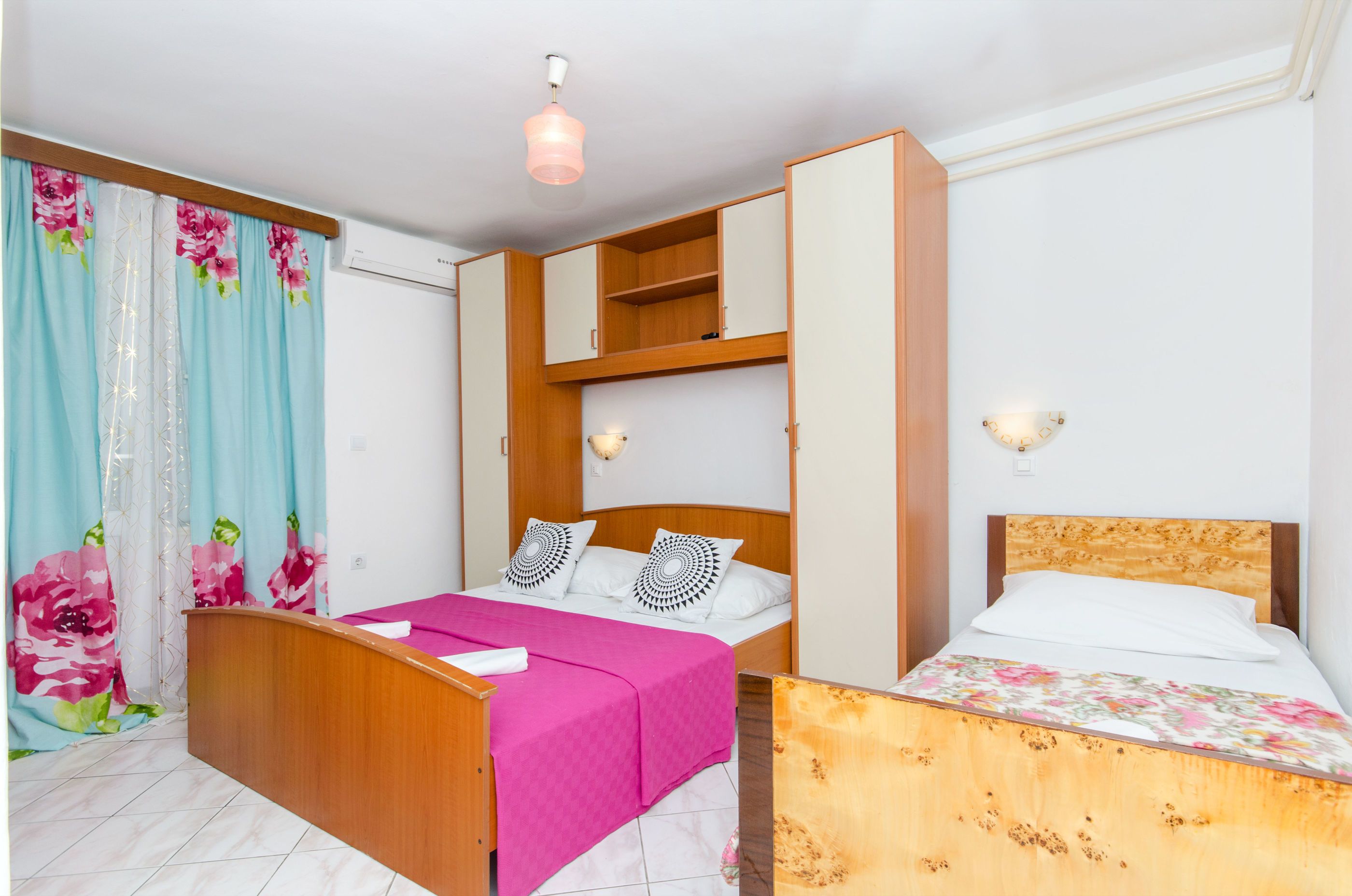 A bedroom with two beds, a wooden wardrobe, a ceiling fan, and a window with curtains.