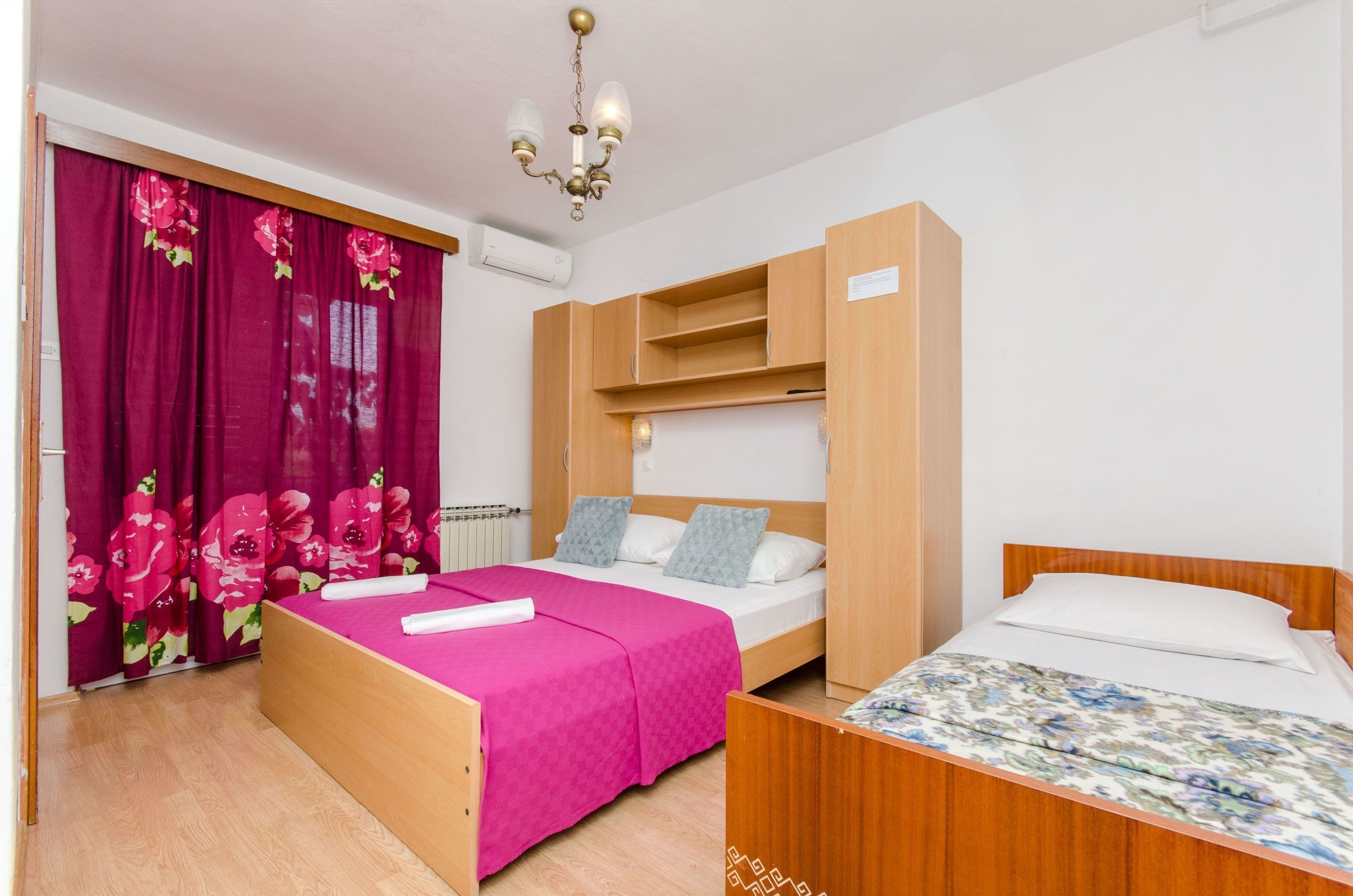 A room with two beds, a wooden wardrobe, a pink bedspread, and a floral bedspread.