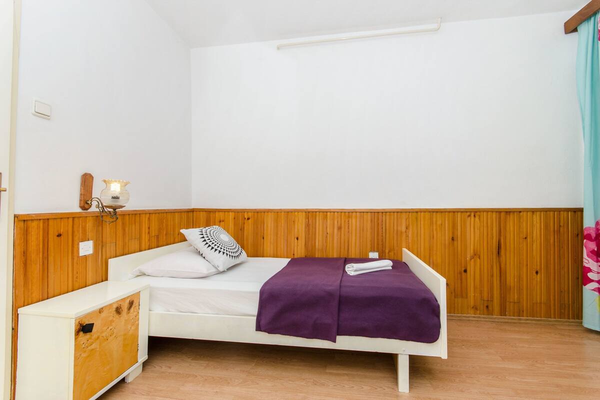Ein Zimmer mit einem Einzelbett, einem Holzkopfstück, einem Nachttisch, einer Wandlampe und einem Fenster mit Vorhang.