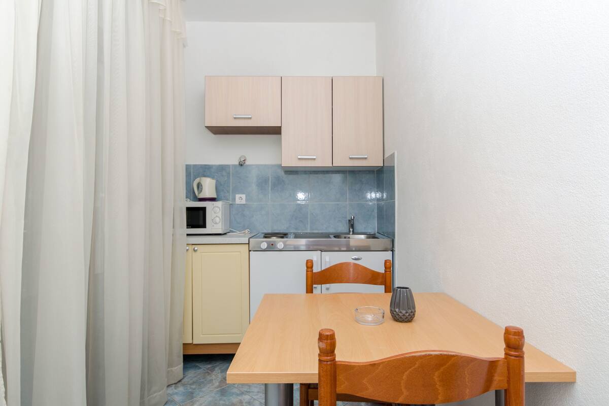 Eine kleine Küche mit Mikrowelle, Herd und Spüle. Ein Holztisch mit Stühlen und einem Glas steht daneben.