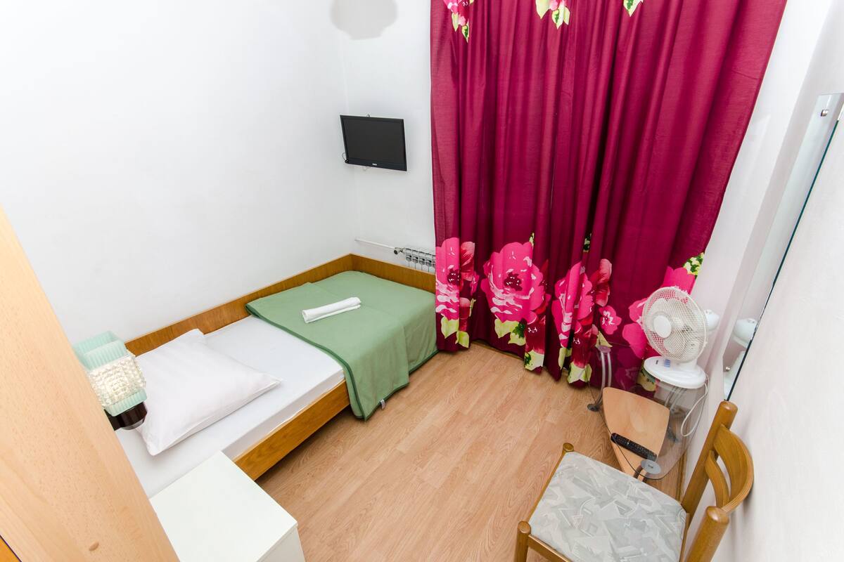 Ein kleines, ordentlich eingerichtetes Schlafzimmer mit einem Einzelbett, einem Nachttisch, einem Stuhl und einem Ventilator.