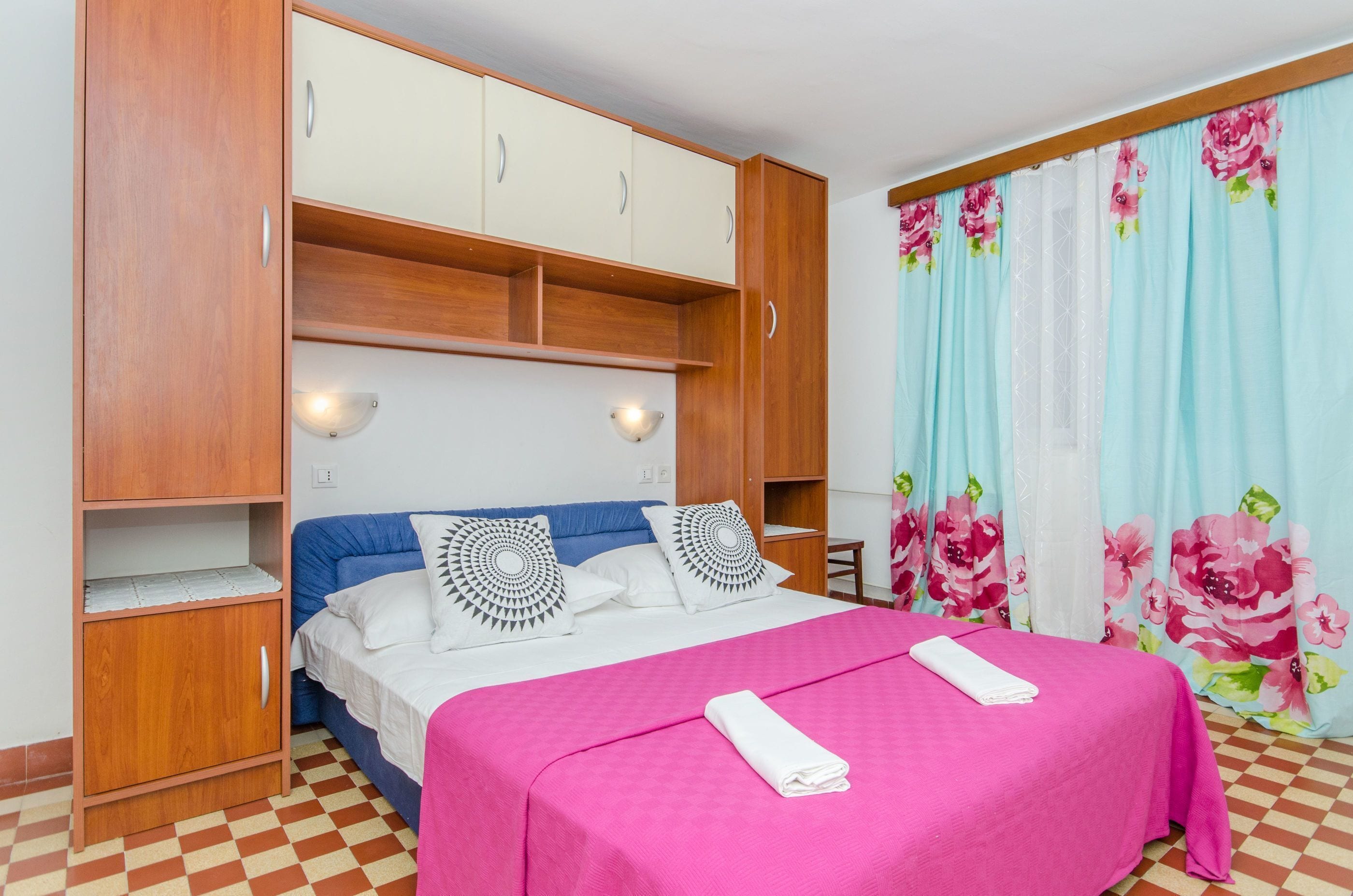 A bedroom with a bed, a wooden wardrobe, a nightstand, and a window with floral curtains.