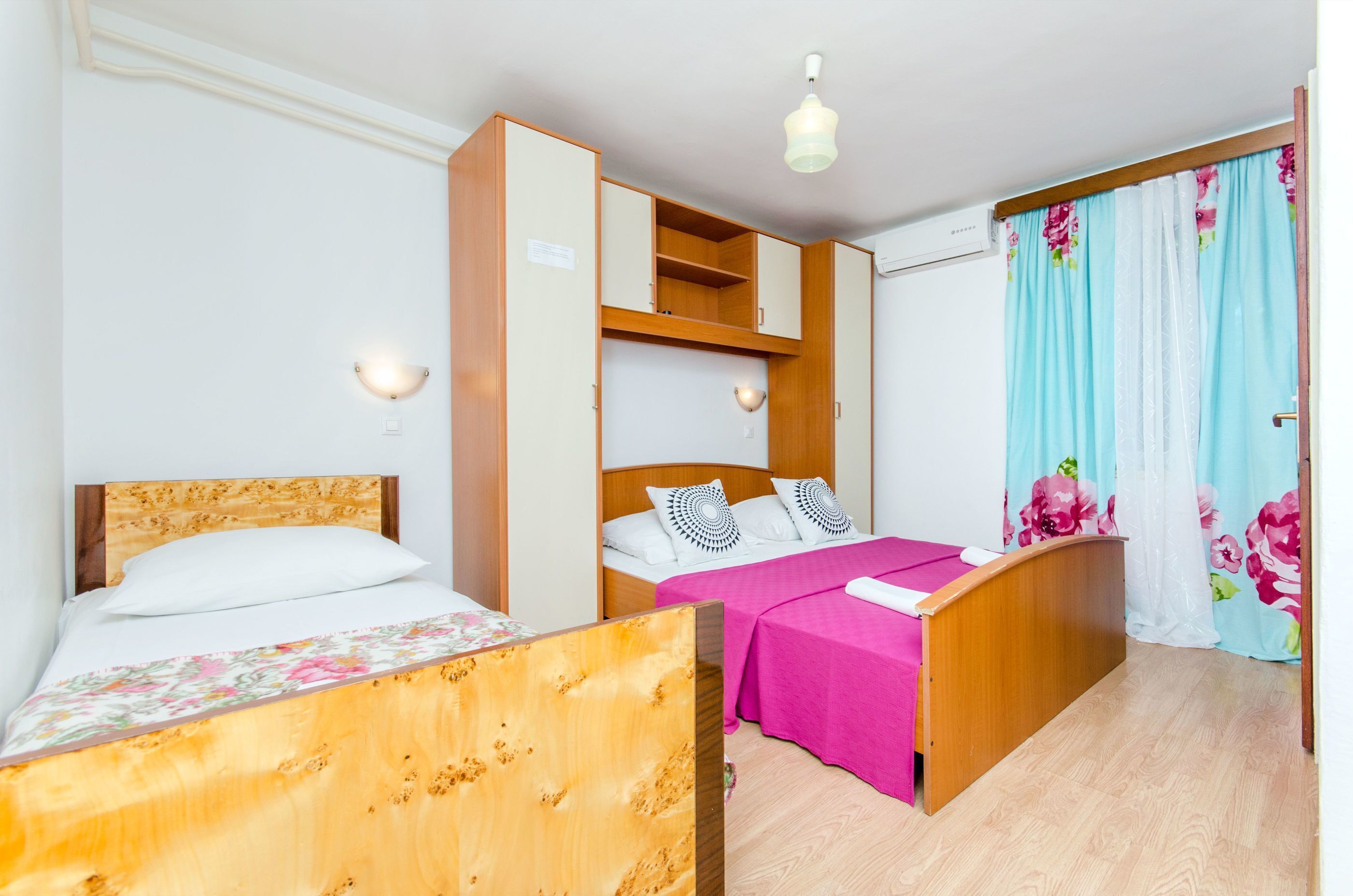 Un dormitorio con dos camas, un armario de madera, una lámpara en el techo y una ventana con cortinas.