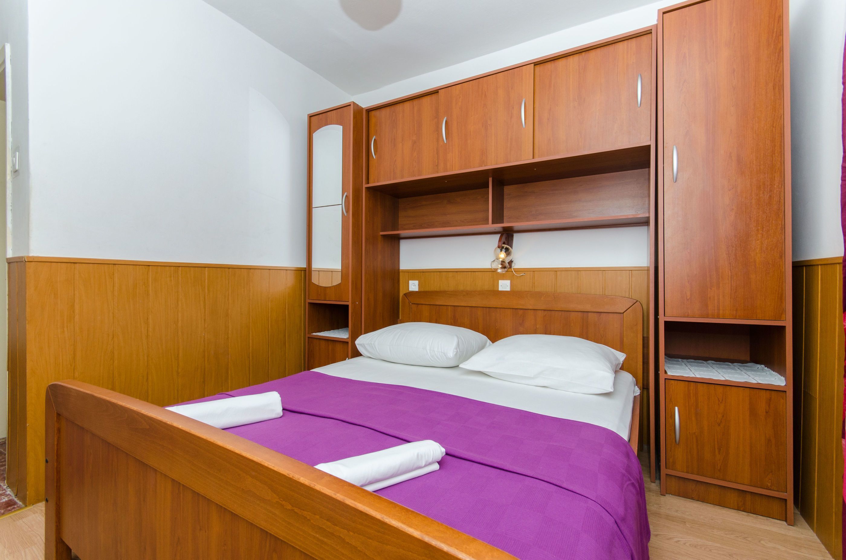 Una cama bien hecha con colcha morada y almohadas blancas, cabecera de madera y armario empotrado de madera con puertas de espejo.