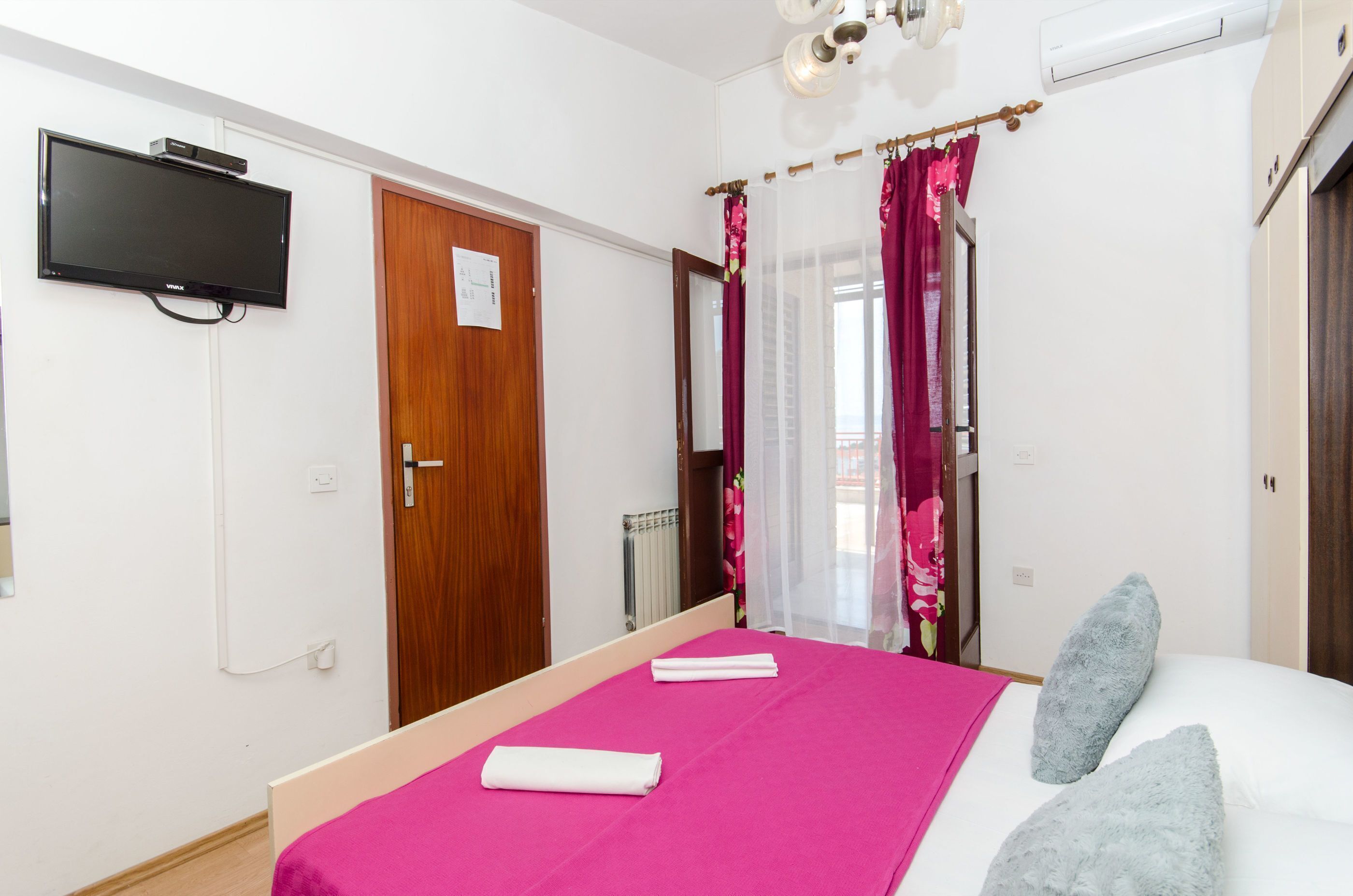 A bedroom with a bed, a flat-screen TV mounted on the wall, a wooden door, and a window with curtains.