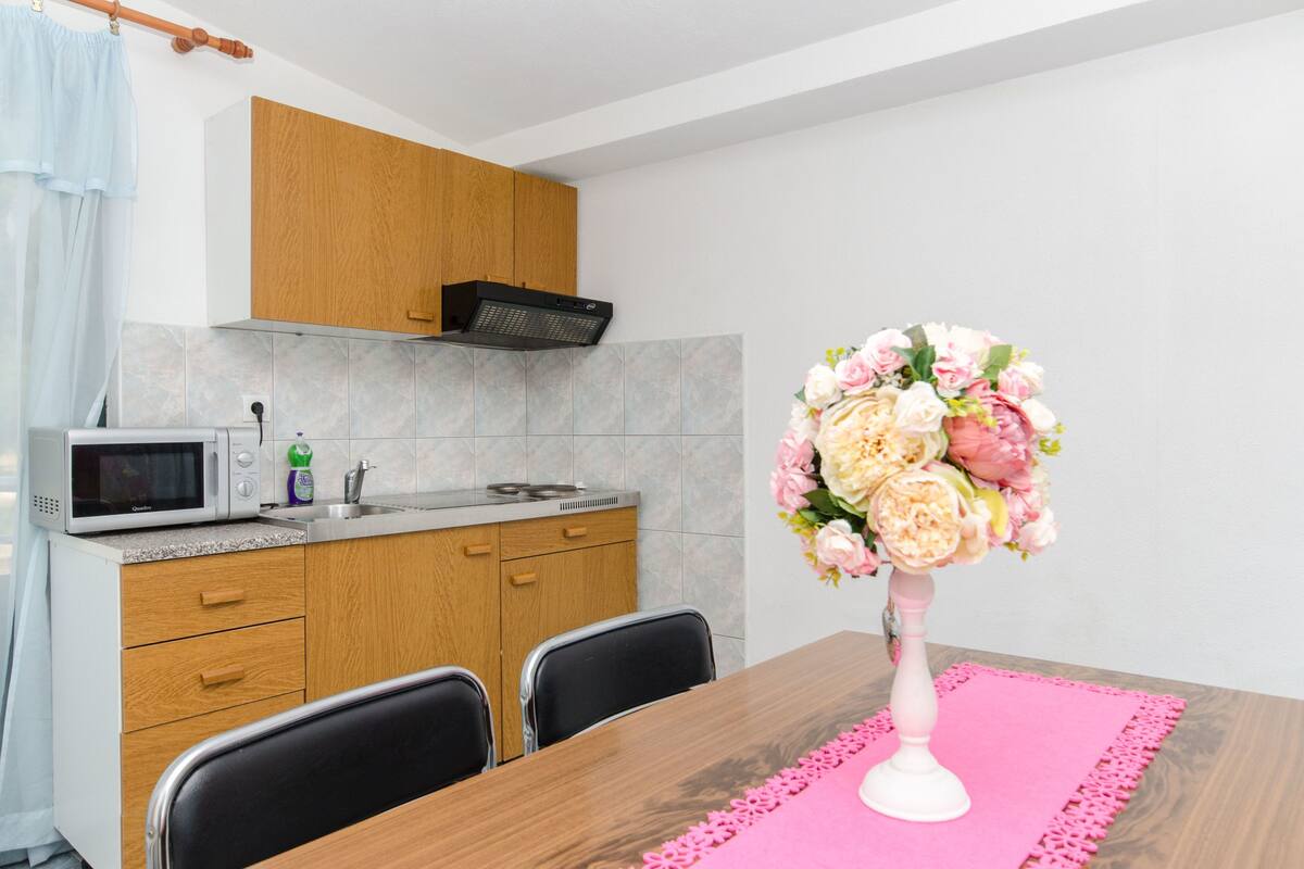 Eine moderne Küche mit Mikrowelle, Holzschränken und einem Esstisch mit Platzdeckchen in Rosa und einer floralen Tischdekoration.