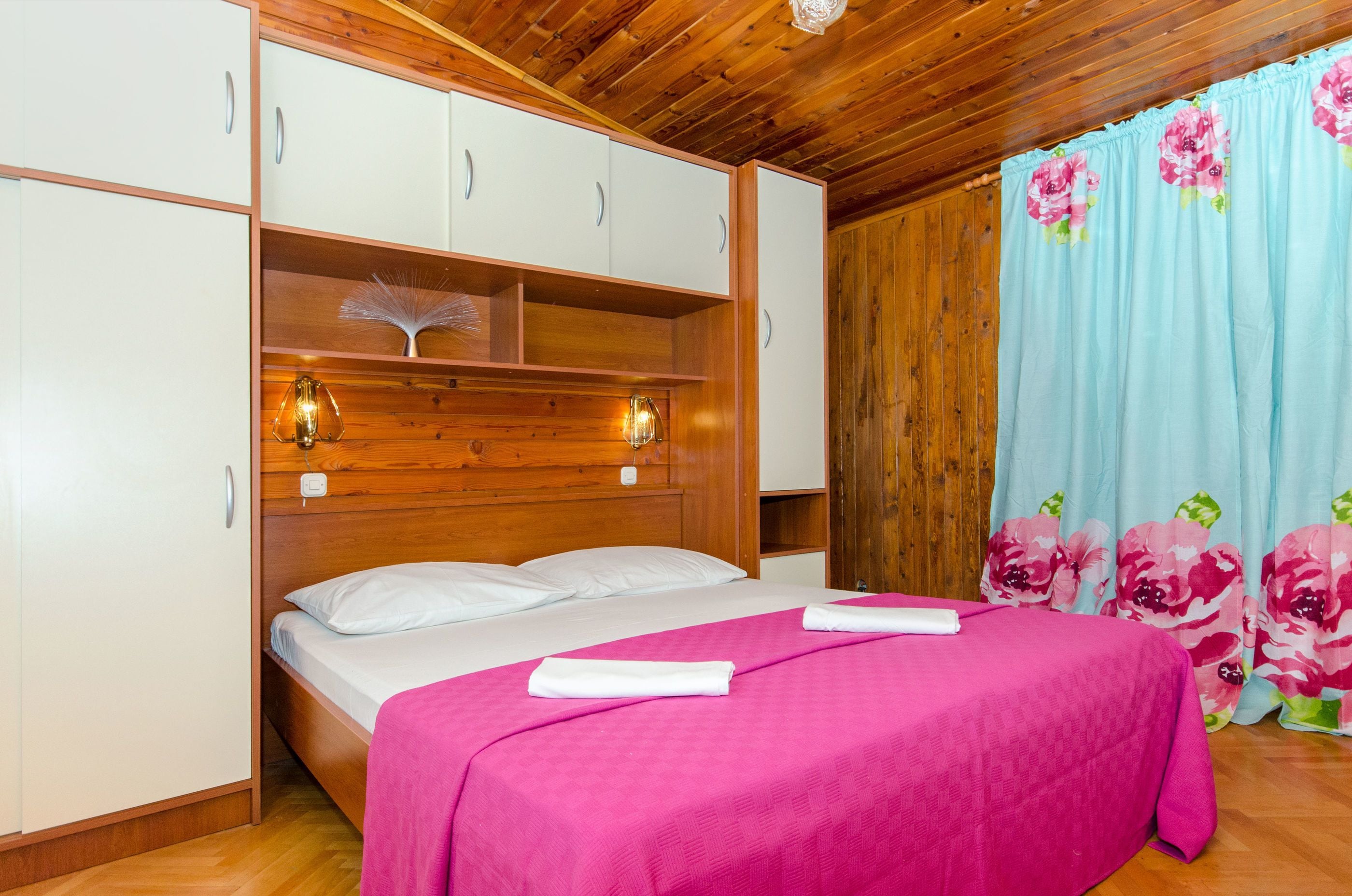Un dormitorio con cabecera de madera, armarios blancos, cama con ropa de cama rosa y cortinas florales.