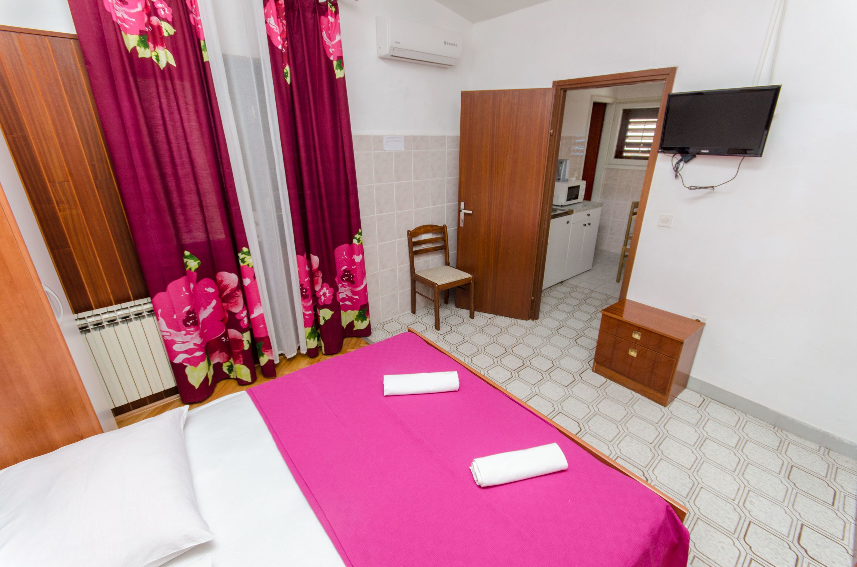 A hotel room with a bed, a wooden nightstand, a chair, a television, and a bathroom visible through an open door.