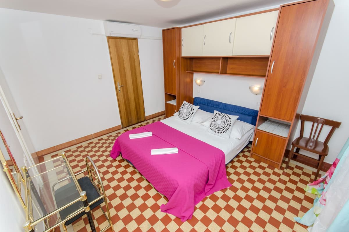 Ein Schlafzimmer mit Schachbrettfußboden, einem Bett mit rosa Bettdecke, einem Holzschrank und einem kleinen Tisch mit Stuhl.