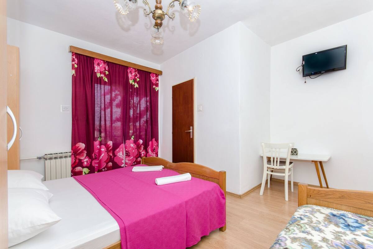 Ein Schlafzimmer mit einem Bett, einem Schreibtisch, einem Sessel, einem Fernseher und einem Fenster mit Vorhängen.