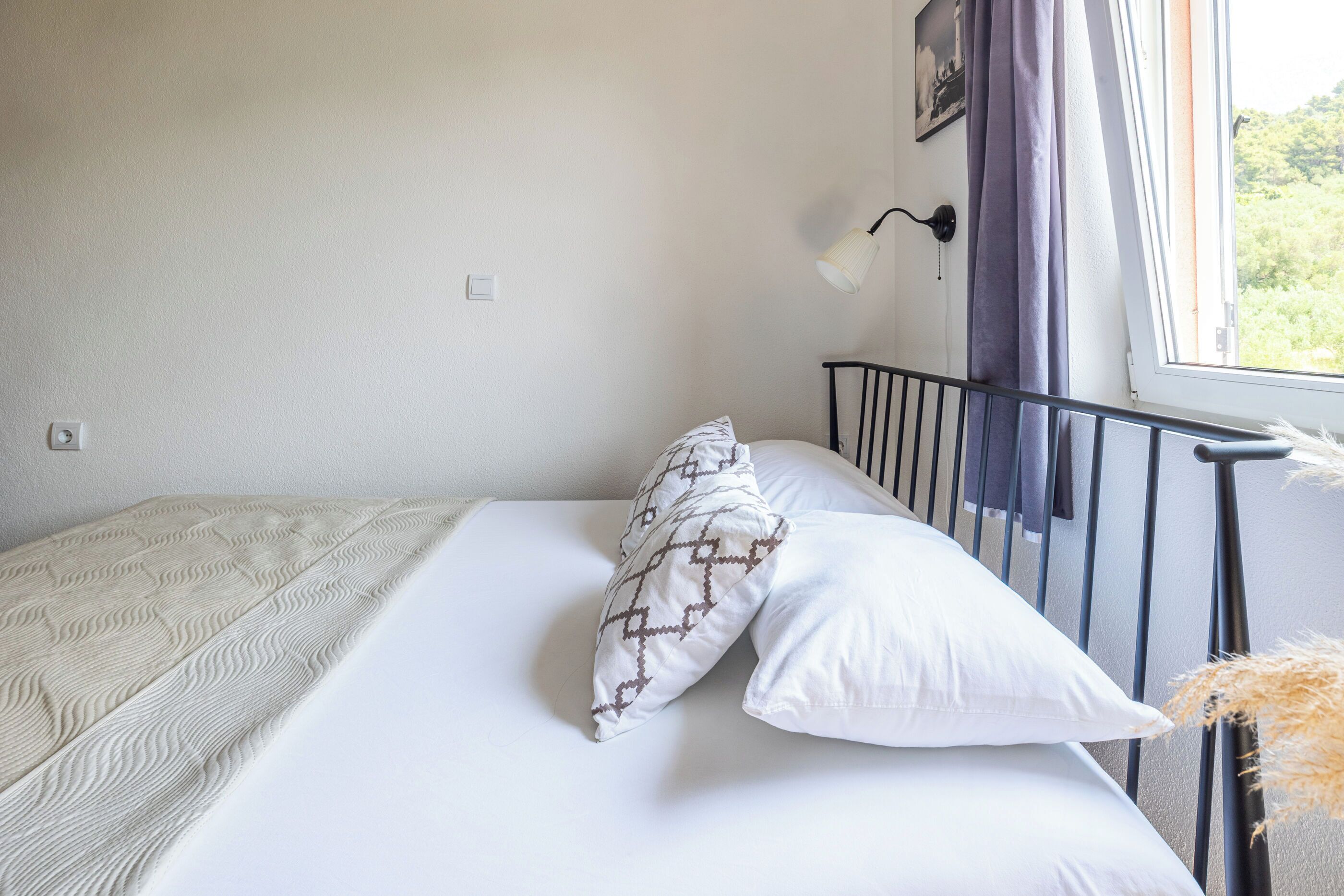 Una cama con ropa de cama blanca y almohadas estampadas, un cabecero de metal negro, una lámpara fijada a la pared y una ventana con vistas a zonas verdes.