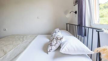 Una cama con ropa de cama blanca y almohadas estampadas, un cabecero de metal negro, una lámpara fijada a la pared y una ventana con vistas a zonas verdes.