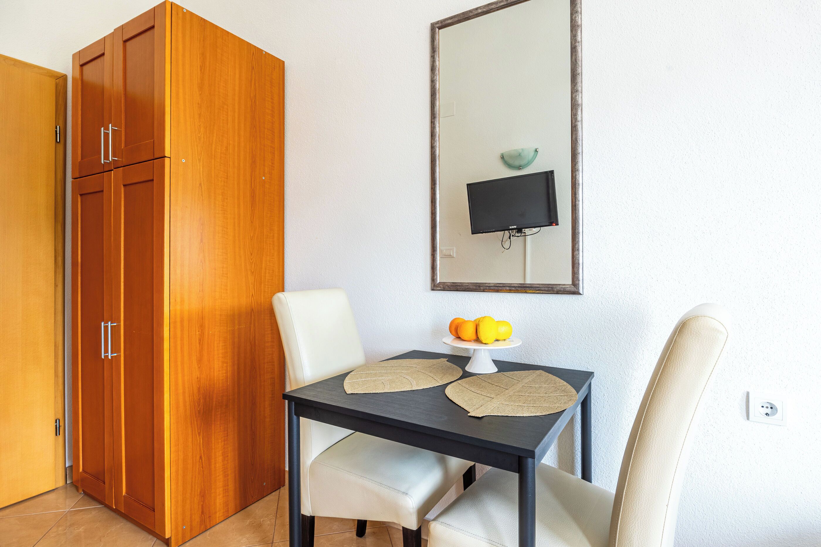 A room with a wooden wardrobe, a dining table with two chairs, a small table with a fruit bowl, and a wall-mounted TV.