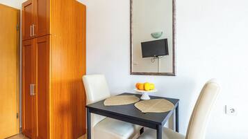 A room with a wooden wardrobe, a dining table with two chairs, a small table with a fruit bowl, and a wall-mounted TV.
