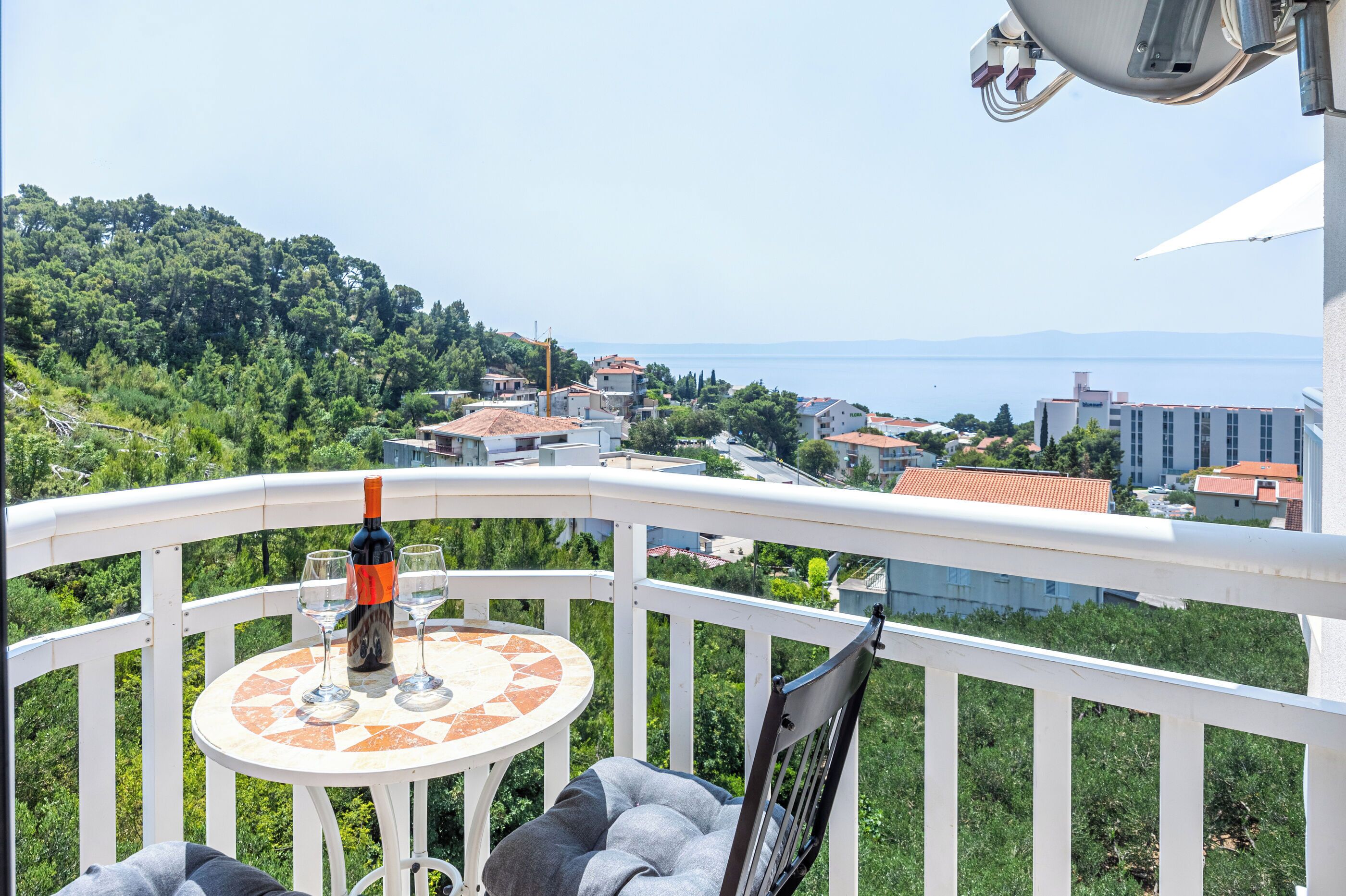 A balcony with a table, a bottle of wine, and glasses, overlooking a town with buildings and a hill.