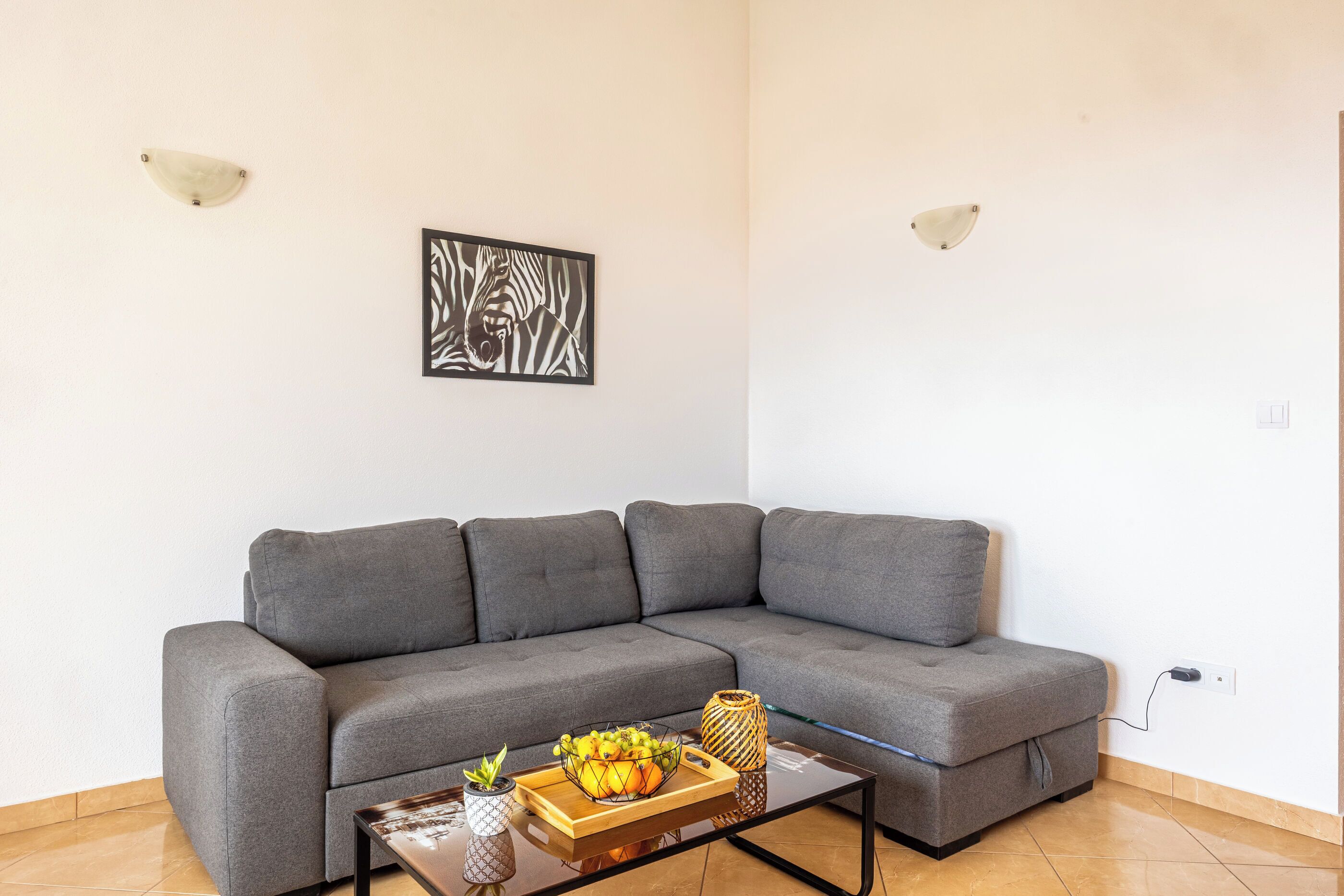 A modern living room with a gray sectional sofa, a glass coffee table with fruit and a vase, and a zebra-patterned artwork on the wall.
