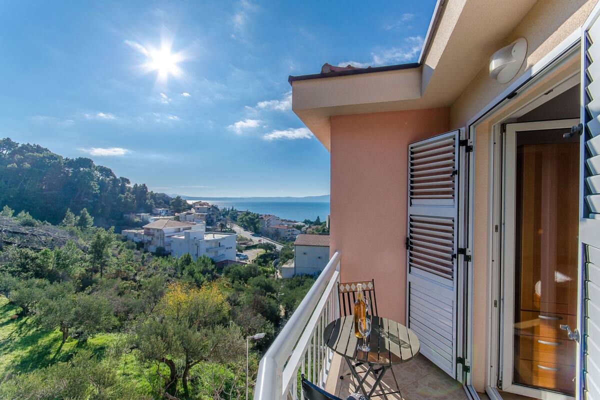 A balcony with a table and chairs, overlooking a coastal town and the sea.