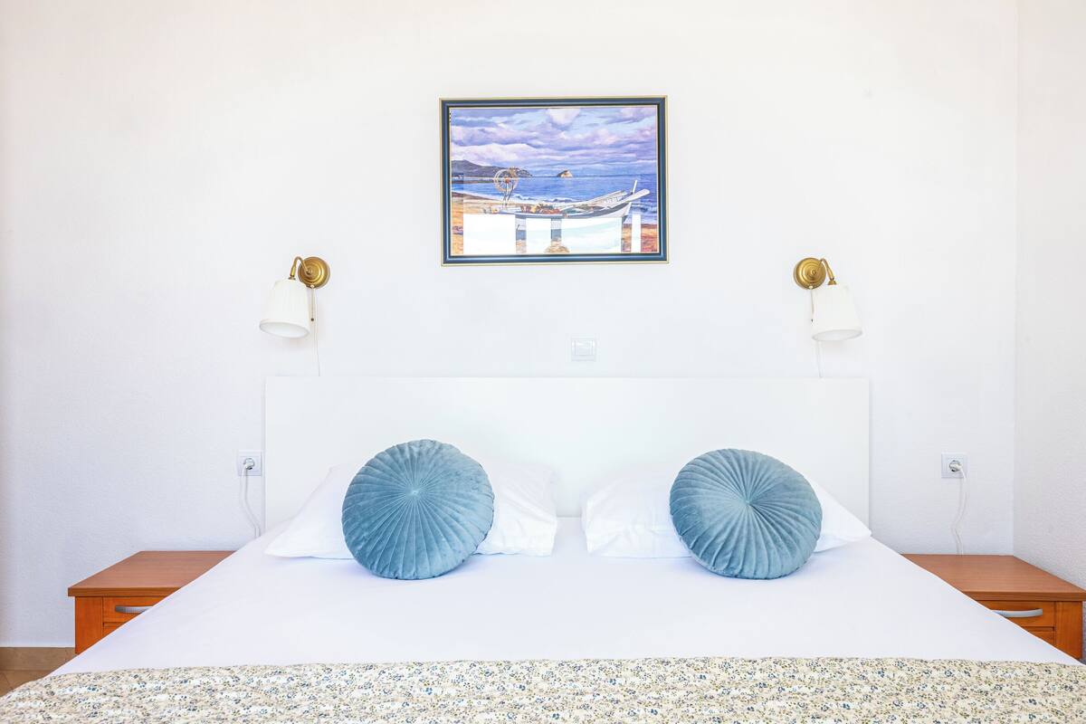 A neatly made bed with two pillows, a framed painting of a boat on a beach, and two wall-mounted lamps.