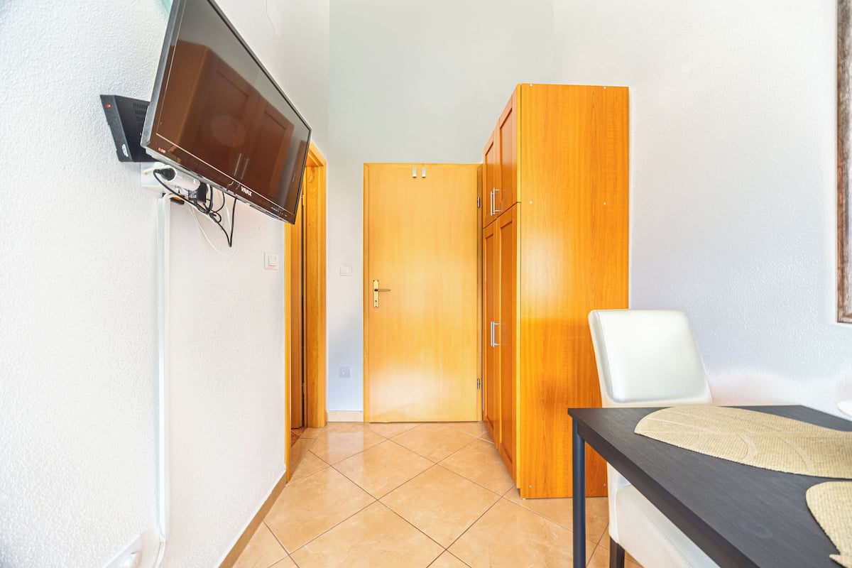 A room with a television mounted on the wall, a wooden wardrobe, a white chair, and a table with a placemat.