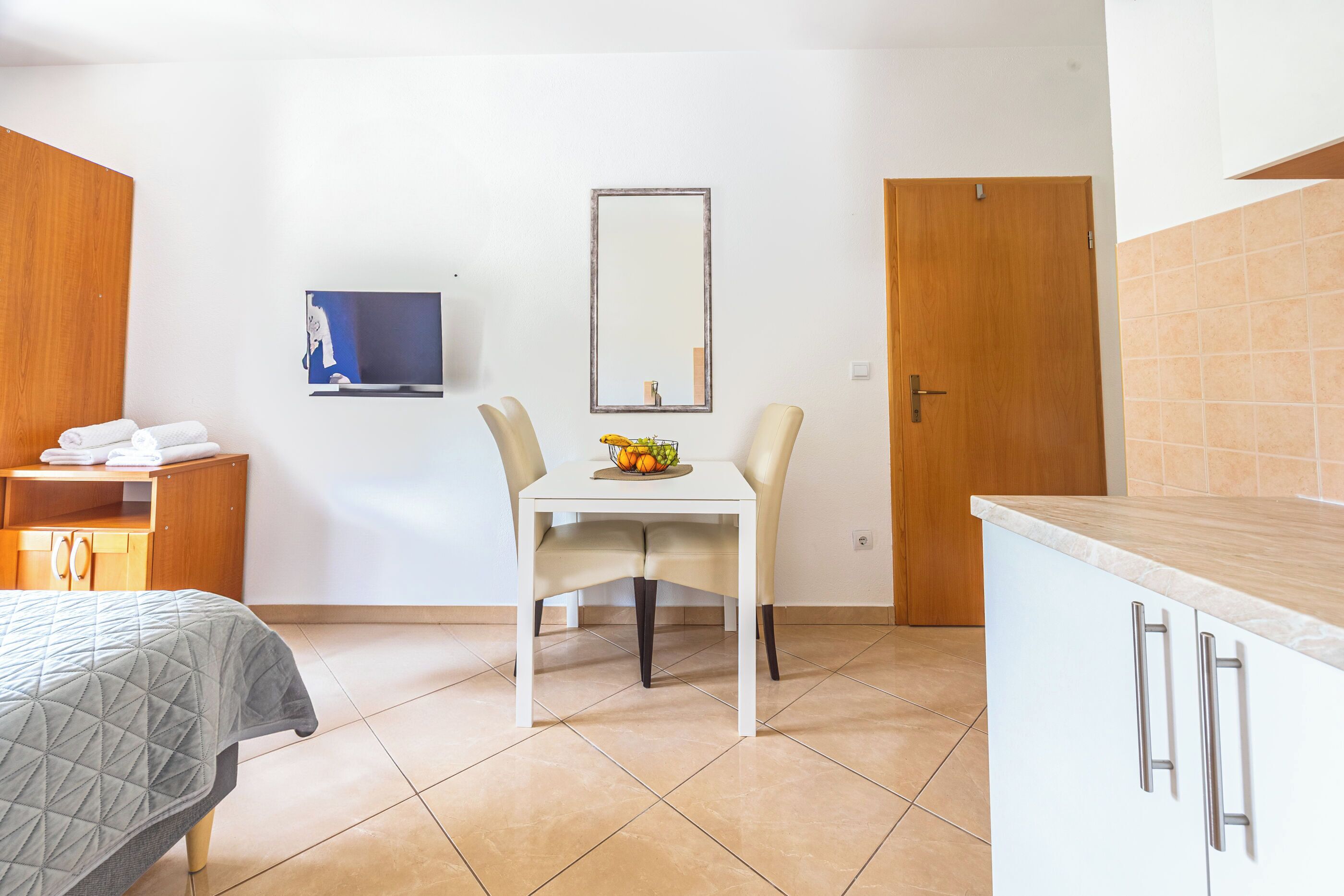 A hotel room with a bed, a small table with chairs, a TV, a wooden door, and a kitchen area.