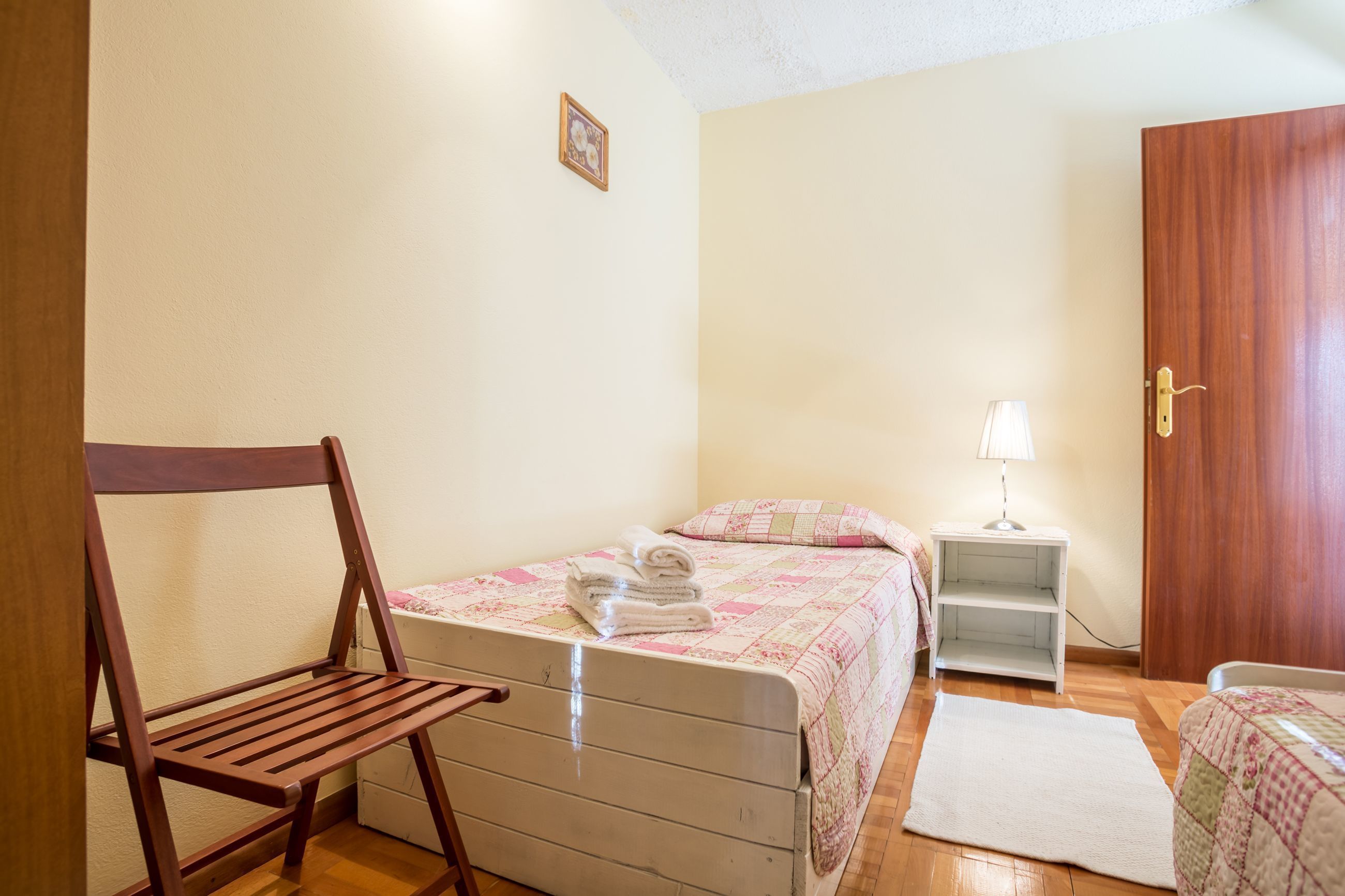 Une chambre à coucher avec un lit, une chaise en bois, une table de chevet avec une lampe et une porte.
