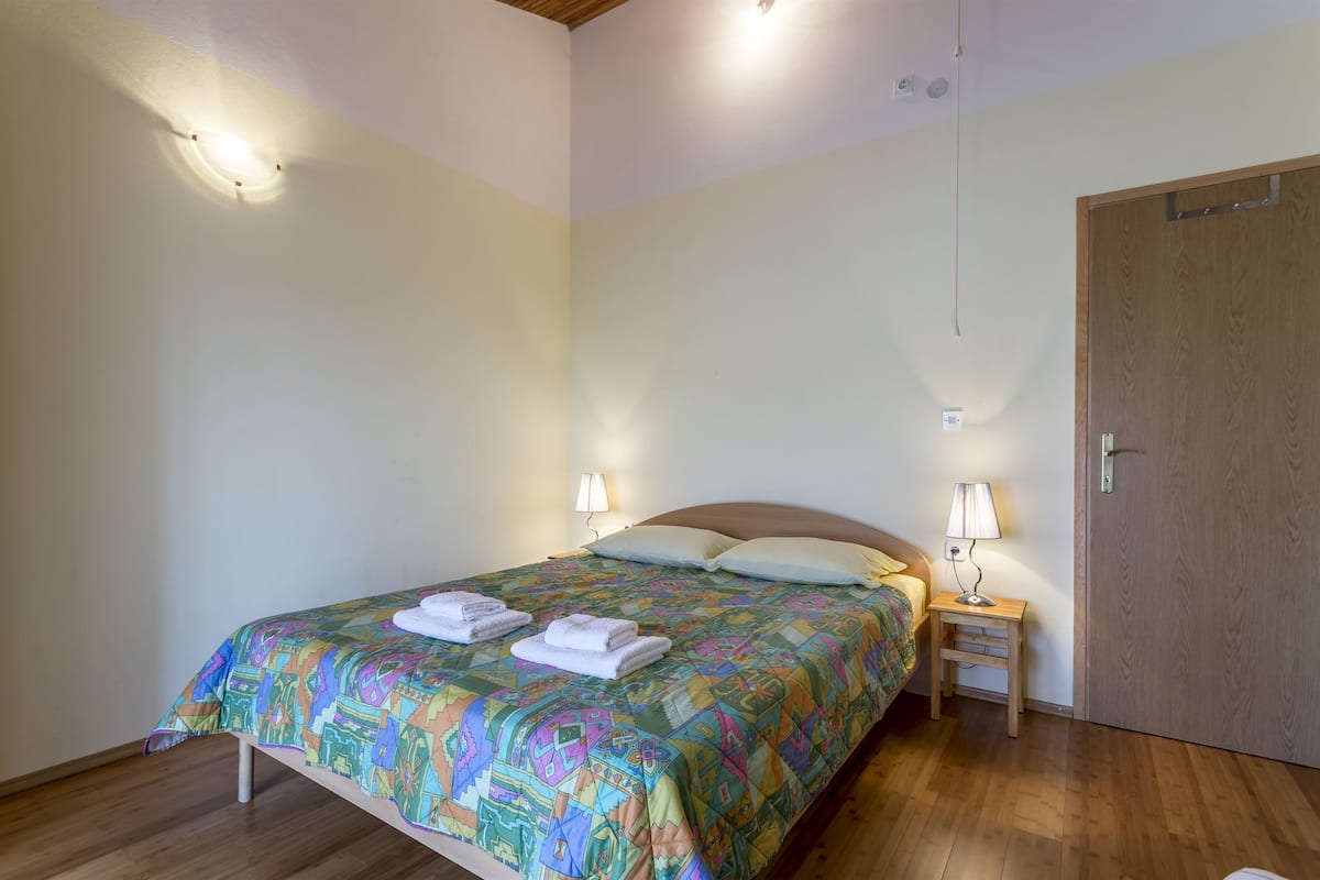 A bedroom with a bed, two bedside lamps, a wooden door, and a patterned bedspread.