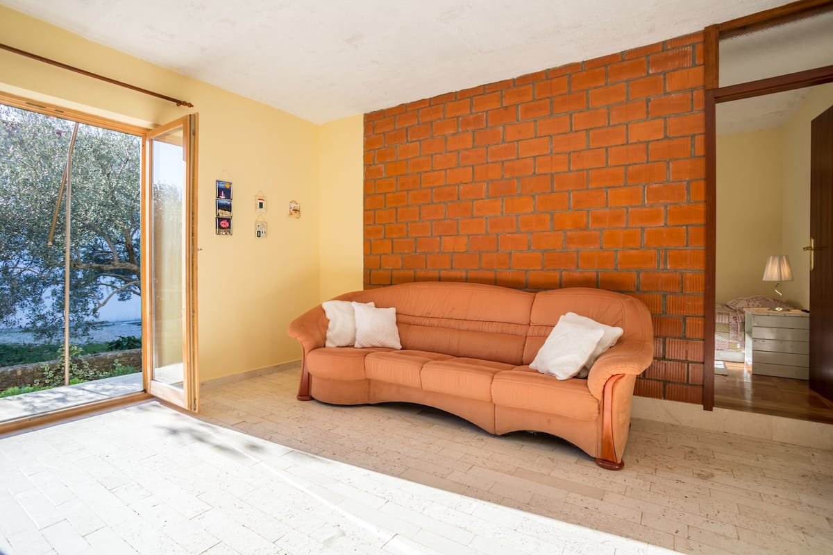 A room with a brick wall, a sofa with pillows, a wooden cabinet, and a glass door leading outside.