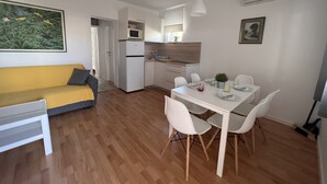 A compact living space with a kitchenette, a dining area with white chairs, a sofa with a yellow cushion, and a painting on the wall. - Apartments Marija Jug (Primosten)