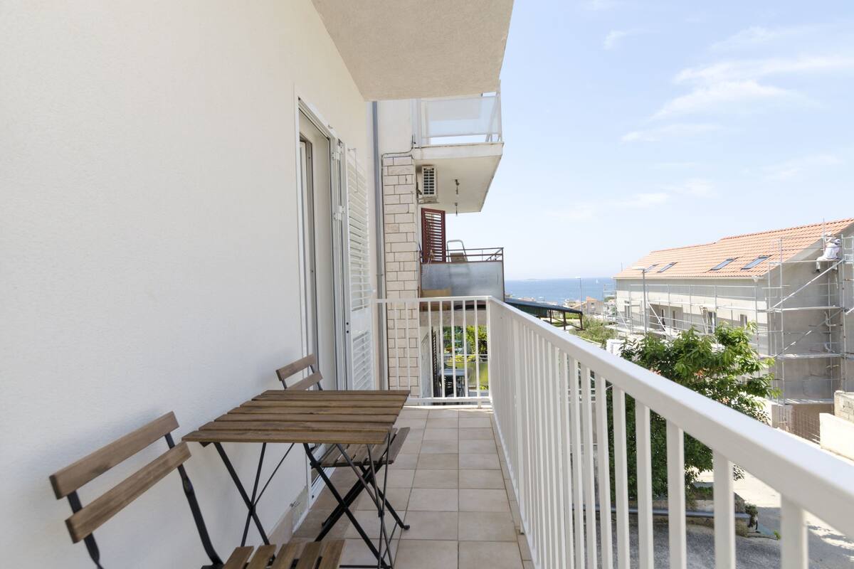 A balcony with a wooden table and chairs, a white railing, and a view of the sea and other buildings.