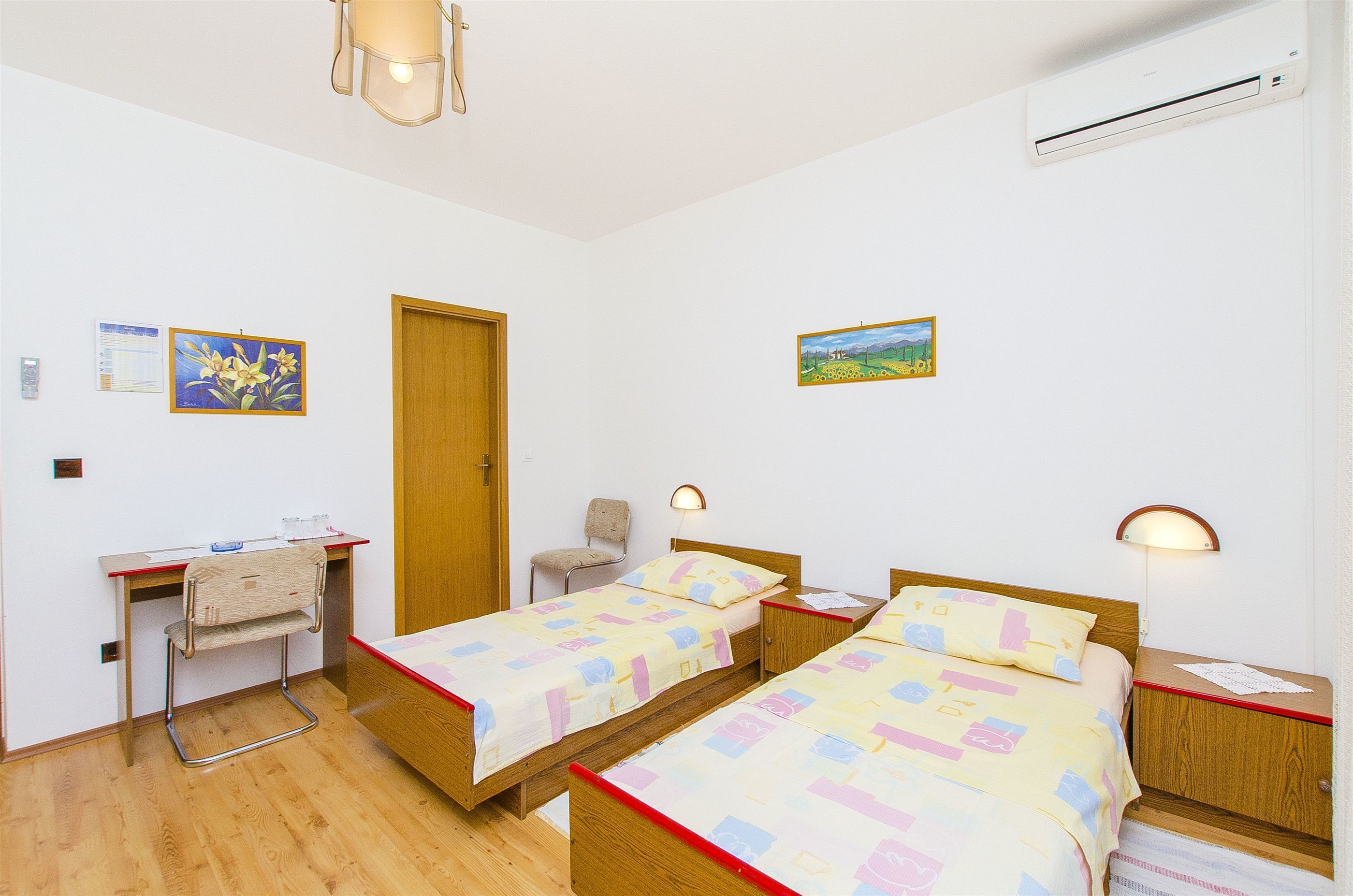 A room with two single beds, a desk with a chair, a wall-mounted air conditioner, and two wall-mounted lamps.
