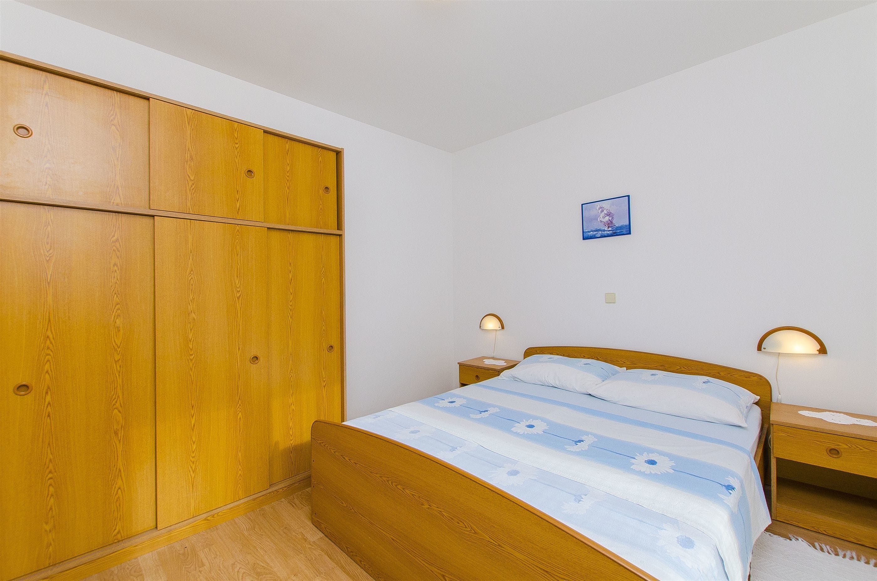 Ein Schlafzimmer mit einem Holzkleiderschrank, einem Bett mit floralem Überwurf, zwei Nachttischen mit Lampen und einem kleinen Bild an der Wand.