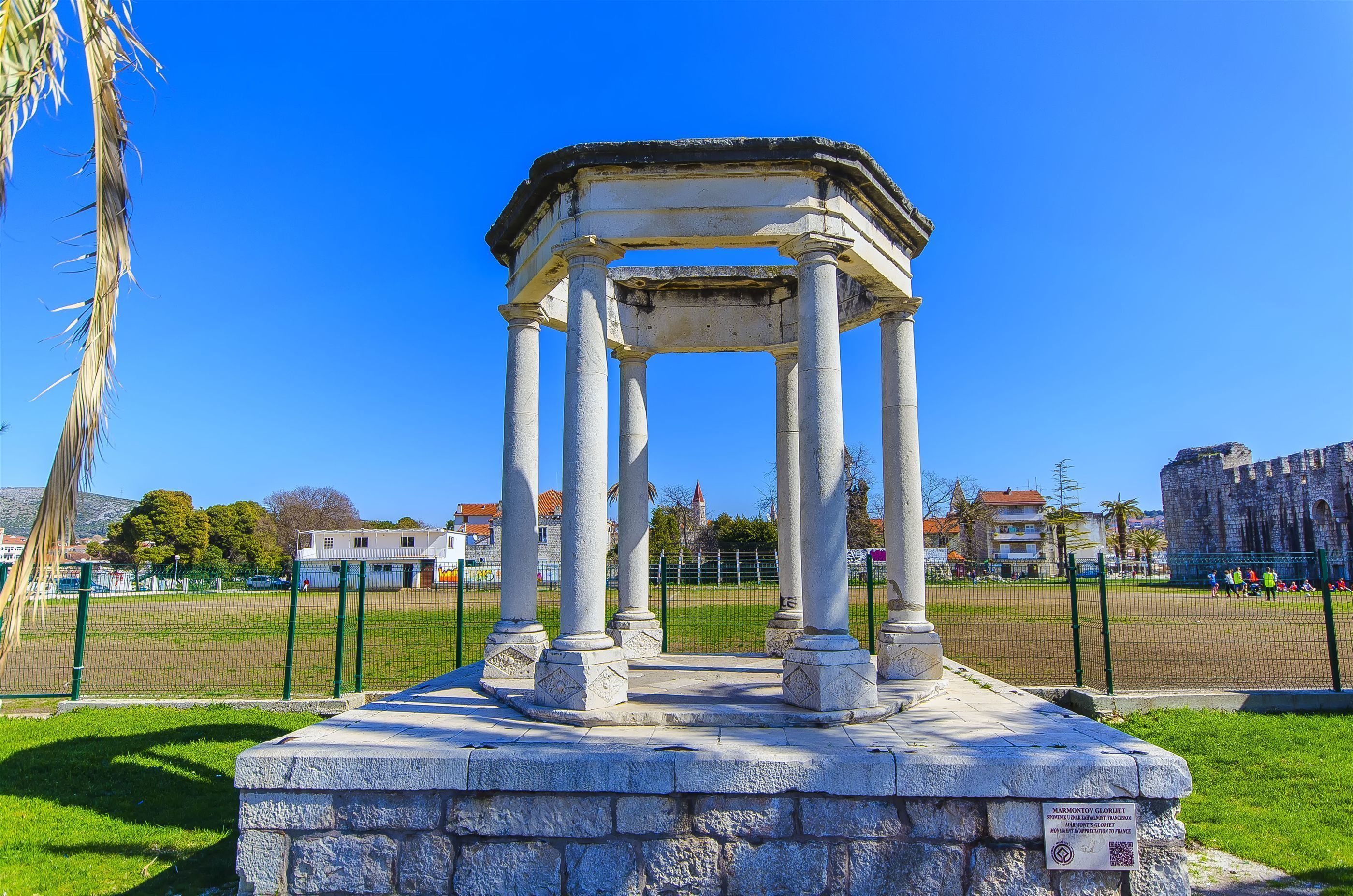 A historical stone structure with columns, surrounded by a grassy area and a fence, with buildings in the background.