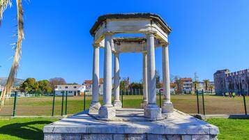 A historical stone structure with columns, surrounded by a grassy area and a fence, with buildings in the background.