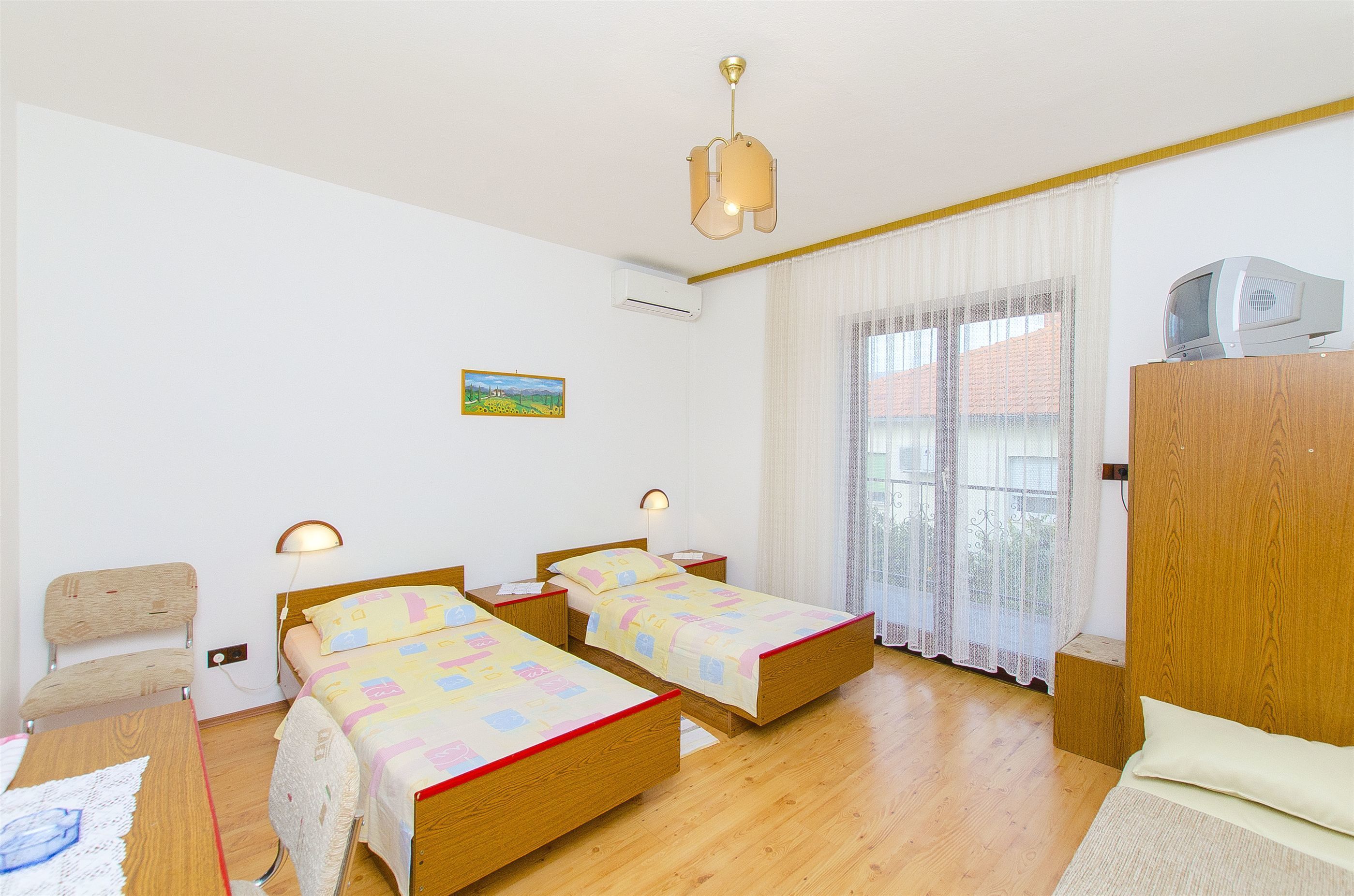 Ein Zimmer mit zwei hölzernen Betten, einem Schreibtisch, einem Stuhl, einem Fernseher auf einer Holzkommode und einem Fenster mit durchsichtigen Vorhängen.